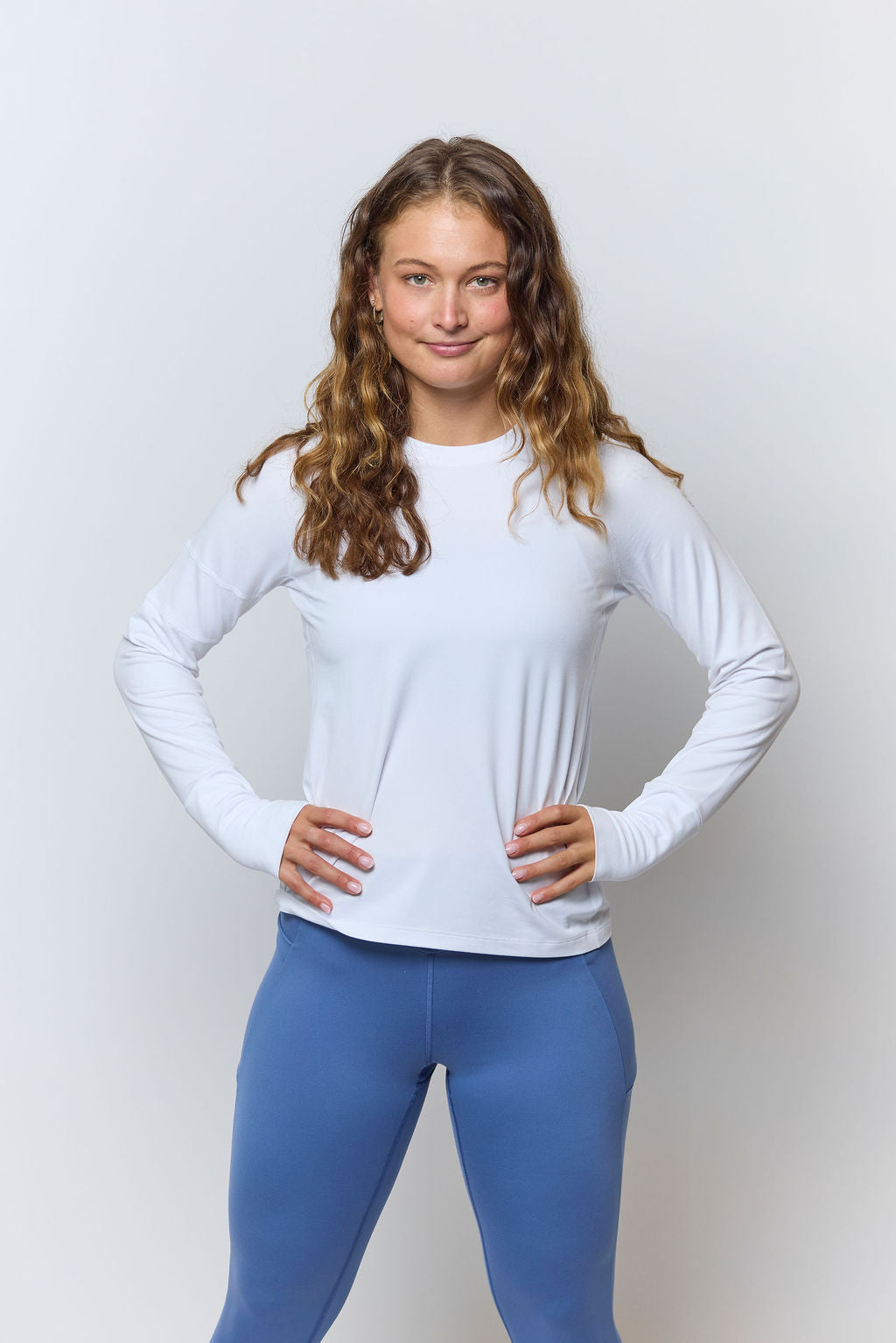 Woman wearing a white long-sleeve top and blue leggings on a light gray background