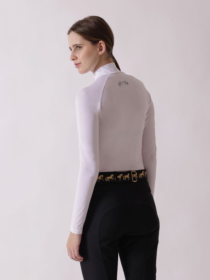 Woman wearing a white long-sleeve top and black riding pants with a gold belt on a light gray background