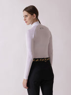 Woman wearing a white long-sleeve top and black riding pants with a gold belt on a light gray background
