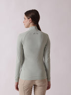 Woman wearing a light green long-sleeve top with a logo on the back against a light pink background
