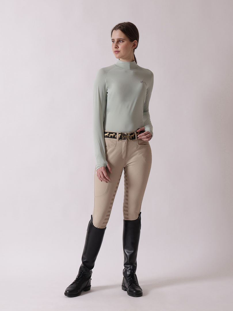 Woman wearing a light green turtleneck sweater, beige riding pants, and black riding boots on a white background