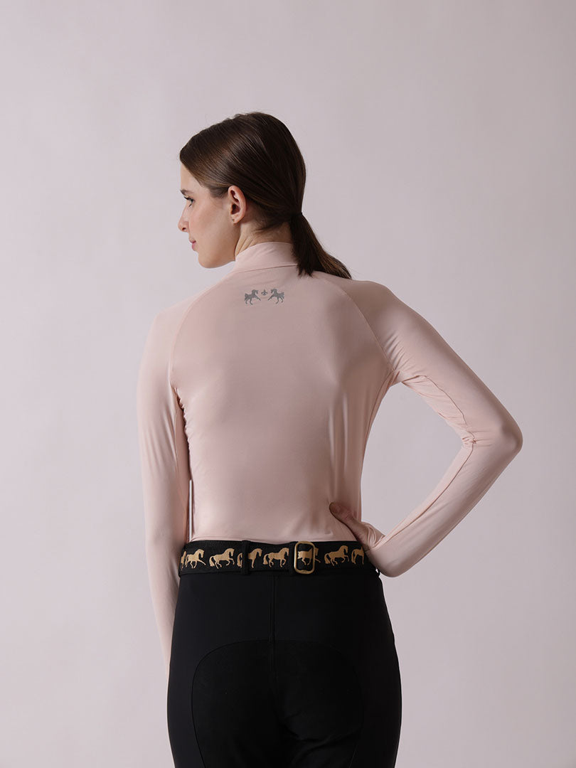 Woman wearing a light pink long-sleeve top and black riding pants with gold horse designs on a plain background