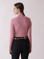 Person wearing a pink long-sleeve top with a logo on the chest and black riding pants with a gold belt on a light pink background.
