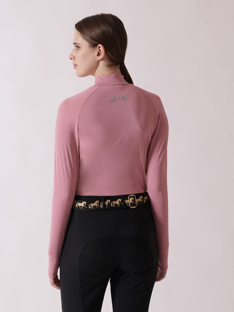 Person wearing a pink long-sleeve top with a logo on the chest and black riding pants with a gold belt on a light pink background.