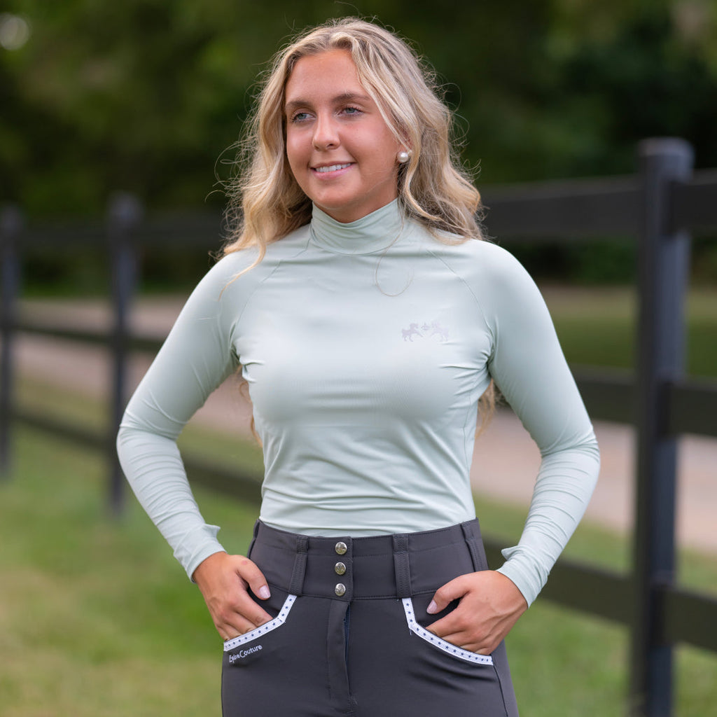 Woman wearing a light green long-sleeve top and dark gray riding pants outdoors.