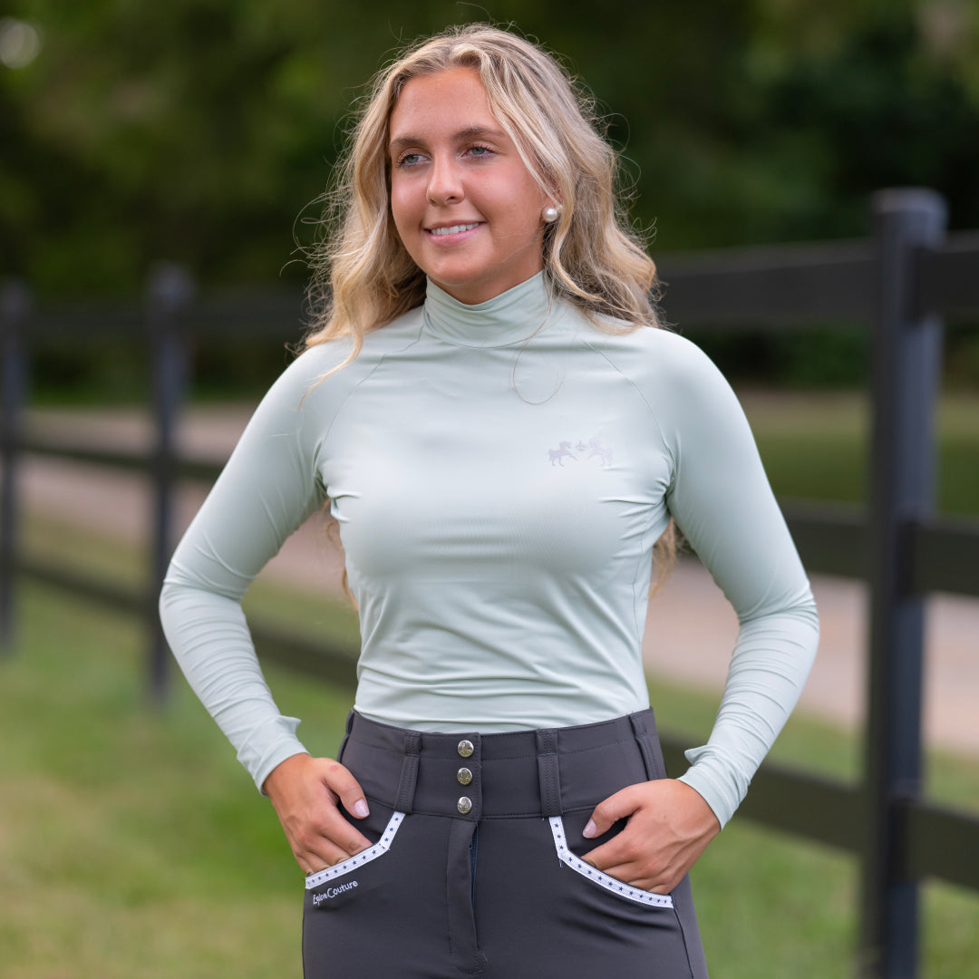 Woman wearing a light green long-sleeve top and dark gray riding pants outdoors.