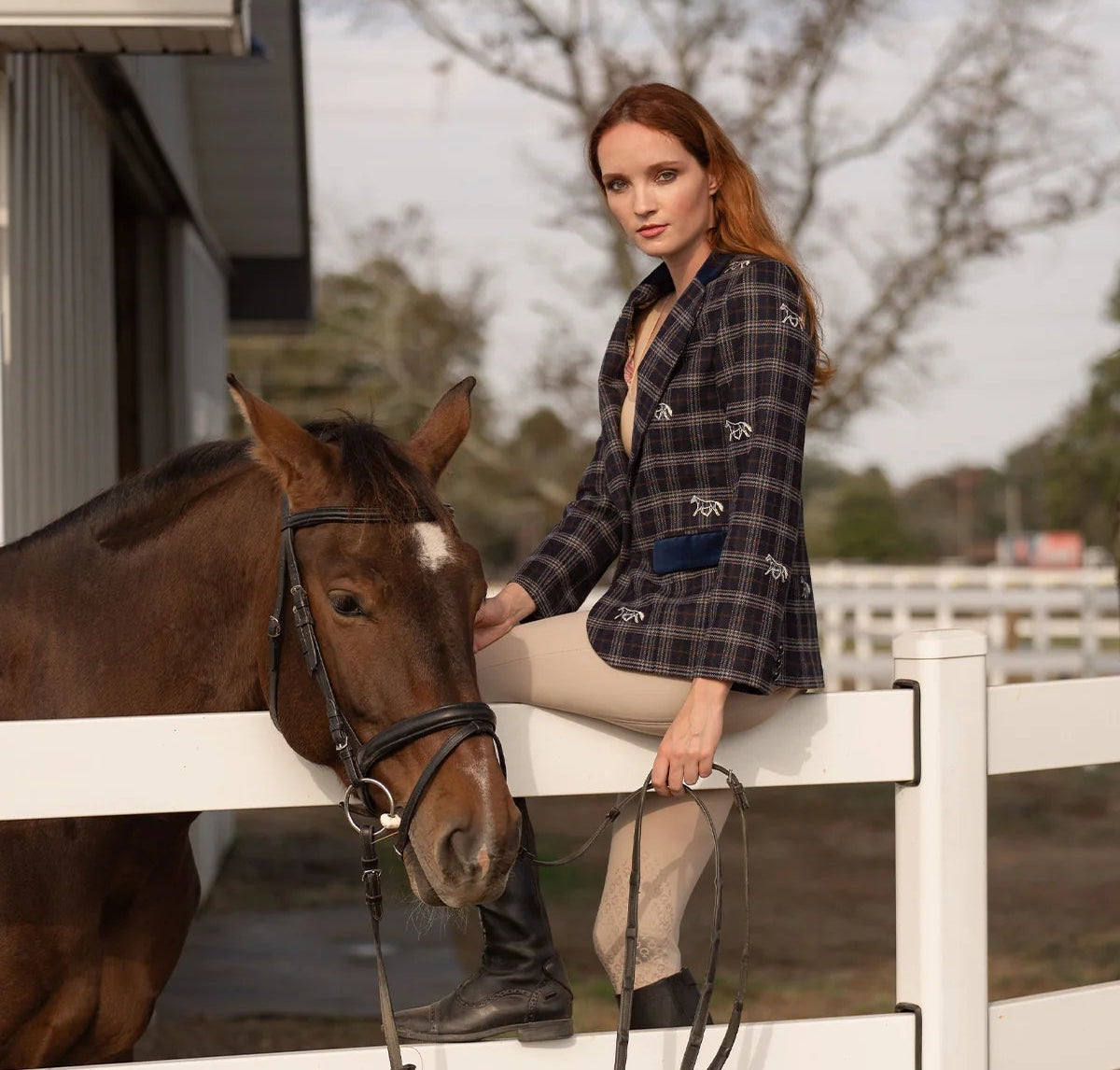 Woman sitting on a white fence with a horse, wearing a plaid shirt and beige pants.