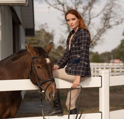 Woman sitting on a white fence with a horse, wearing a plaid shirt and beige pants.
