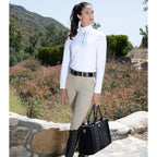 Woman in equestrian attire holding a black bag outdoors with greenery in the background