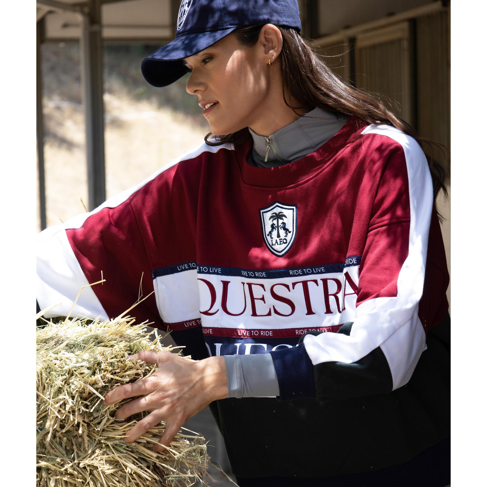 Person wearing a maroon and navy 'Oquestra' sweatshirt with a logo, holding hay.