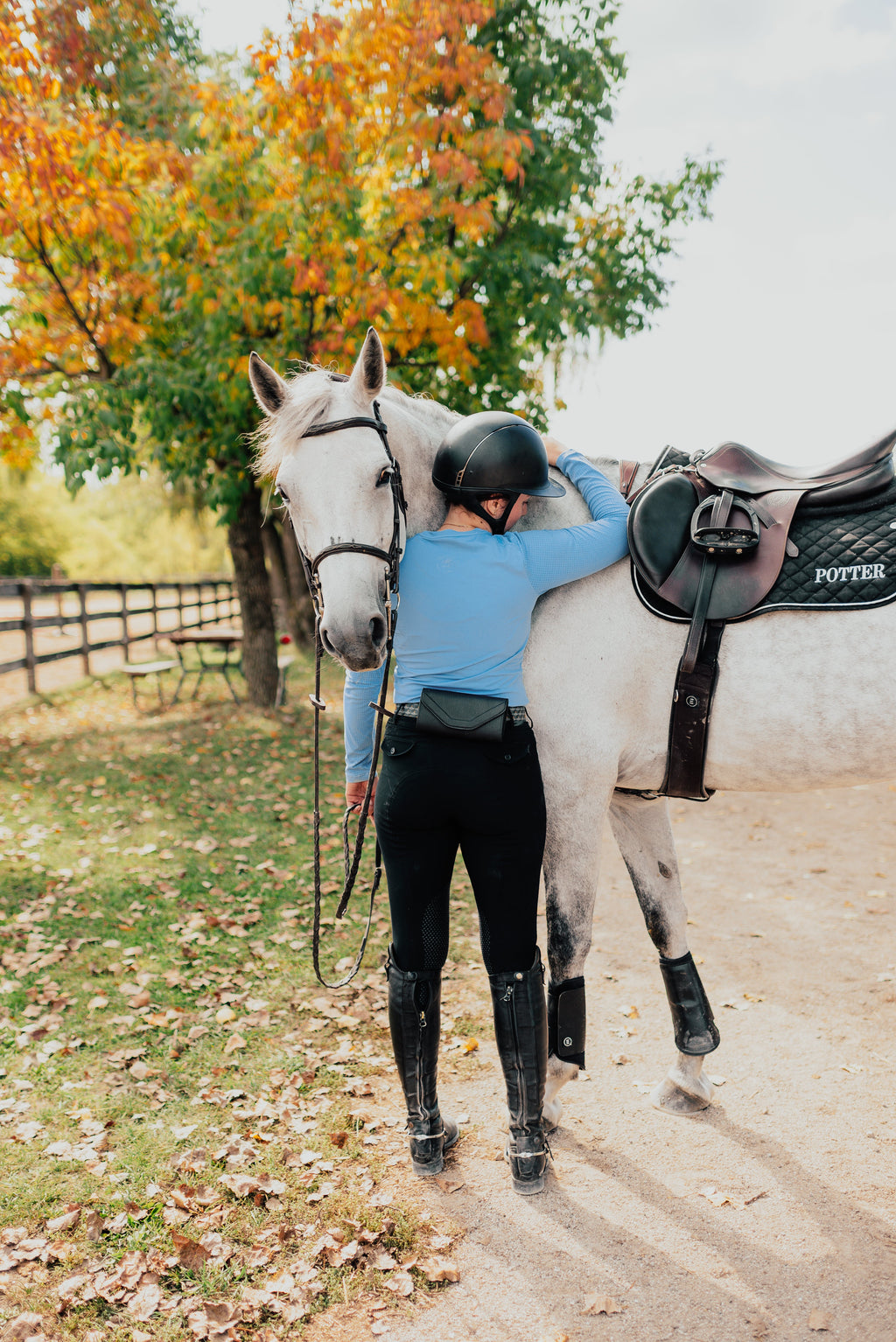 Person in equestrian attire standing next to a white horse on a path with trees in the background.