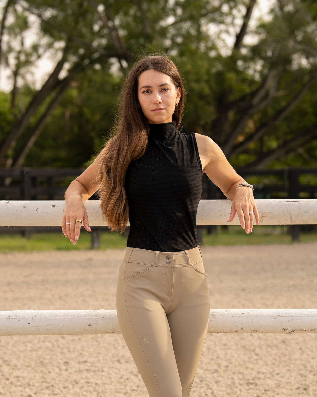 Woman in black top and beige pants standing in an equestrian arena.