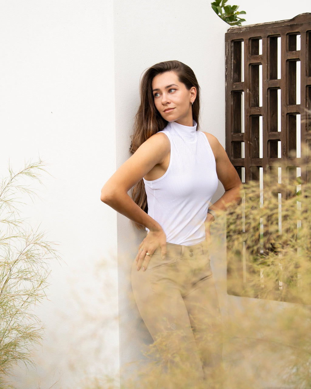 Woman in a white sleeveless top and beige pants standing against a white wall with plants around.