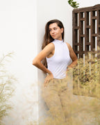 Woman in a white sleeveless top and beige pants standing against a white wall with plants around.