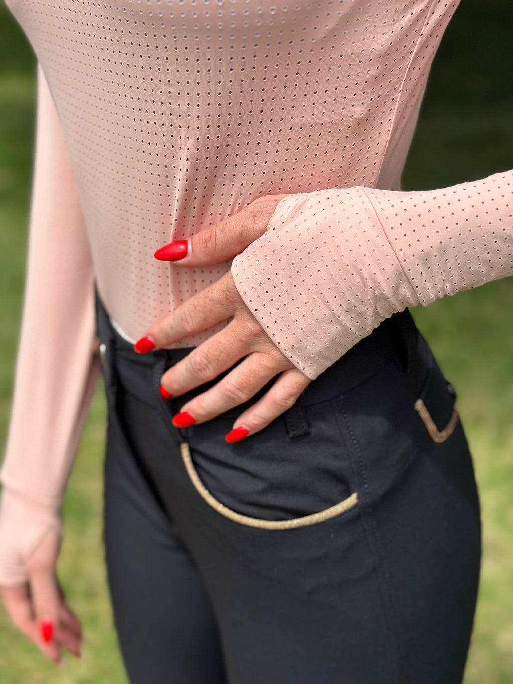Person wearing a light pink long sleeve top and dark riding pants with red nail polish against a blurred green background