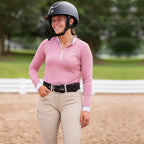 Woman in equestrian attire with pink shirt and beige pants standing on a dirt path.