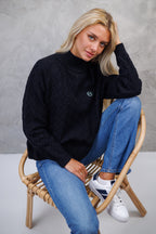 Woman sitting on a chair wearing a black sweater and blue jeans against a gray background
