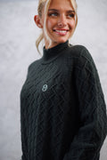 Woman wearing a black cable knit sweater with a logo on a gray background