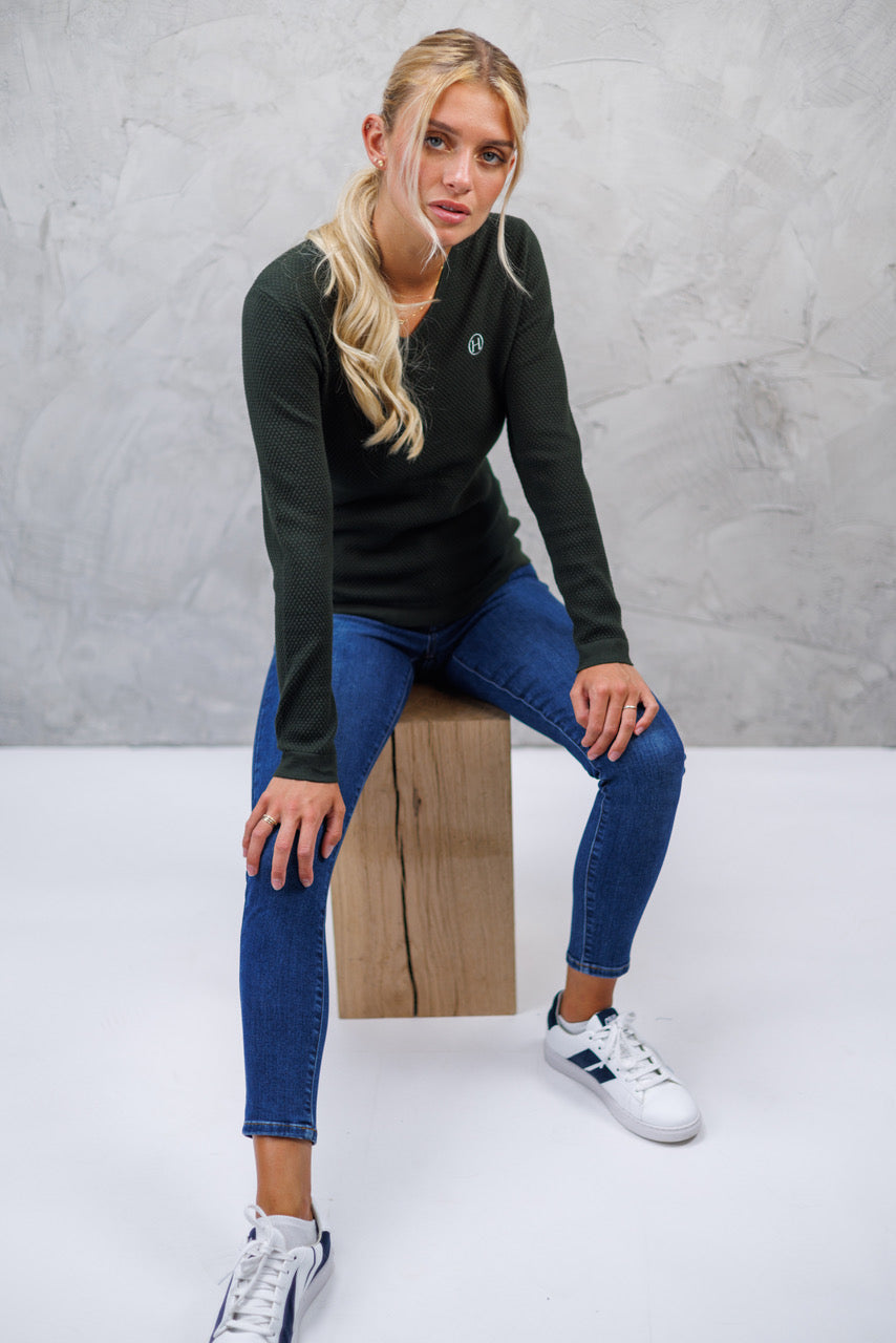 Woman sitting on a wooden block wearing a black long-sleeve shirt and blue jeans against a gray background