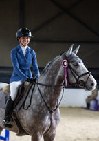 Woman in equestrian attire riding a gray horse with a pink rosette in an indoor arena.