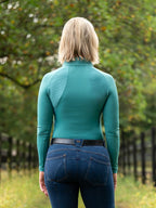 Person wearing a teal long-sleeve top and blue jeans standing in a grassy area with trees.