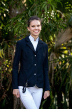 Woman wearing a navy airbag-compatible equestrian jacket and white pants standing in front of green foliage.